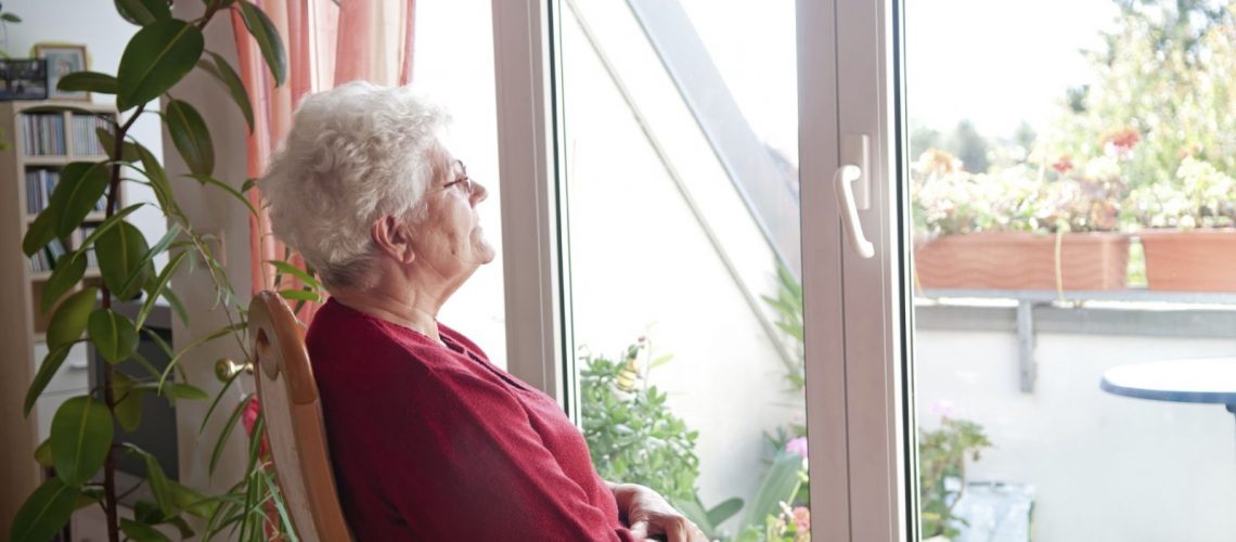 Senior woman in isolation at home looking outside her backyard window.